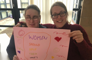 Two GDA members smile, one with her arm around the other, both hold a poster hand decorated which says 'Women should have their voices heard'