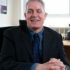 GDA Trustee Professor Bill Hughes, smiling with his hands clasped on his desk, in his office. Bill has short light grey hair and is wearing a blue shirt and tie and a pinstriped suit.