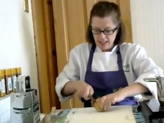A chef conducts an online cookery demonstration. She is stood at a chopping board chopping vegetables in white clothes and a purple apron.