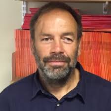 GDA Trustee, Professor Nick Watson. Nick has brown hair and a brown and grey beard, he is wearing a dark polo shirt and stands smiling in front of a shelf full of brightly coloured books.