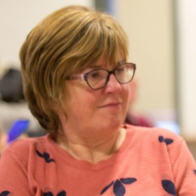 GDA Trustee, Maureen McHugh -.Maureen has a dark blonde bob and glasses, and is looking off camera, in discussion, in a community hall. Maureen is wearing a peach jumper with navy flowers on it.
