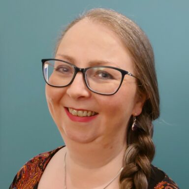 Marianne Scobie smiles at the camera. She is wearing glasses and red lipstick. A plaited braid rests on her left shoulder. She is wearing a necklace with a purple stone.