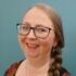 Marianne Scobie smiles at the camera. She is wearing glasses and red lipstick. A plaited braid rests on her left shoulder. She is wearing a necklace with a purple stone.