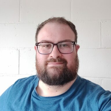 Kevin has short hair, dark rimmed glasses and a full beard. He's wearing a blue tshirt.