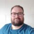Kevin has short hair, dark rimmed glasses and a full beard. He's wearing a blue tshirt.