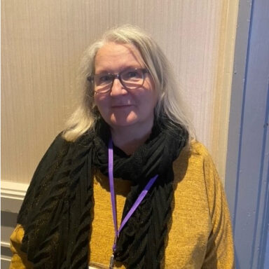 Fiona is wearing a mustard top and green scarf. She has shoulder length hair and glasses.