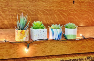 four small plant pots made of jesmonite sit on a piano music shelf with small succulent plants in them and fairy lights draped across. the jesmonite is white with speckles and streaks of colour from the repurposed plastics added.