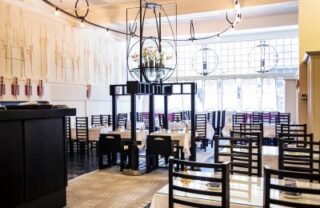 The Willow Tearooms in the afternoon, with light coming through the windows. High backed Rennie Mackintosh chairs are sat at tables of 4 with purple Mackintosh designs on the walls.