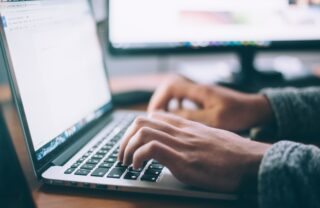 a close up of a silver laptop open on a wooden surface, the photograph focuses on a person's hands typing, illuminated by the screen.