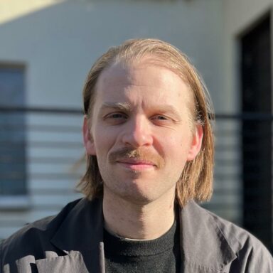 Photo of Lewis Watson. He has chin-length hair and a moustache.