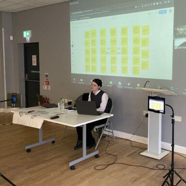 Meg sits at a table, with a Zoom screen projected on the wall behind her, showing digital sticky notes with text on them. Next to her is an iPad.