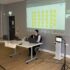 Meg sits at a table, with a Zoom screen projected on the wall behind her, showing digital sticky notes with text on them. Next to her is an iPad.