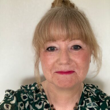 Close-up photo of Angela looking at the camera and smiling. She has blonde hair, a fringe, and is wearing pink lipstick. Her shirt is patterned in black and green colours.