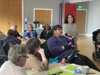 Paula speaks into a microphone at a GDA event. Other GDA members at staff are watching and listening.