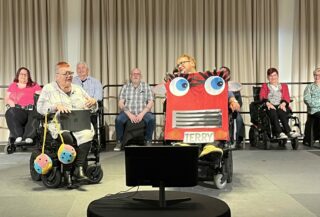 The Purple Poncho Players perform, with Paula and Rose at the front of the stage. Paula is wearing a large red car costume, with big eyes and eyelashes on the front, and a licence plate reading 'TERRY'. Rose has a large pair of fluffy dice hanging off her wheelchair.