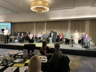 The Purple Poncho Players perform on stage. They are raising their fists triumphantly. Paula is at the front.
