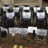 Empty wheelchairs and pairs of shoes are grouped together. Signs in the wheelchairs and behind the shoes read signs like 'I was depressed', 'I cost too much', 'I was coerced', 'I will be next', 'I had no support', 'I wasn't valued'.
